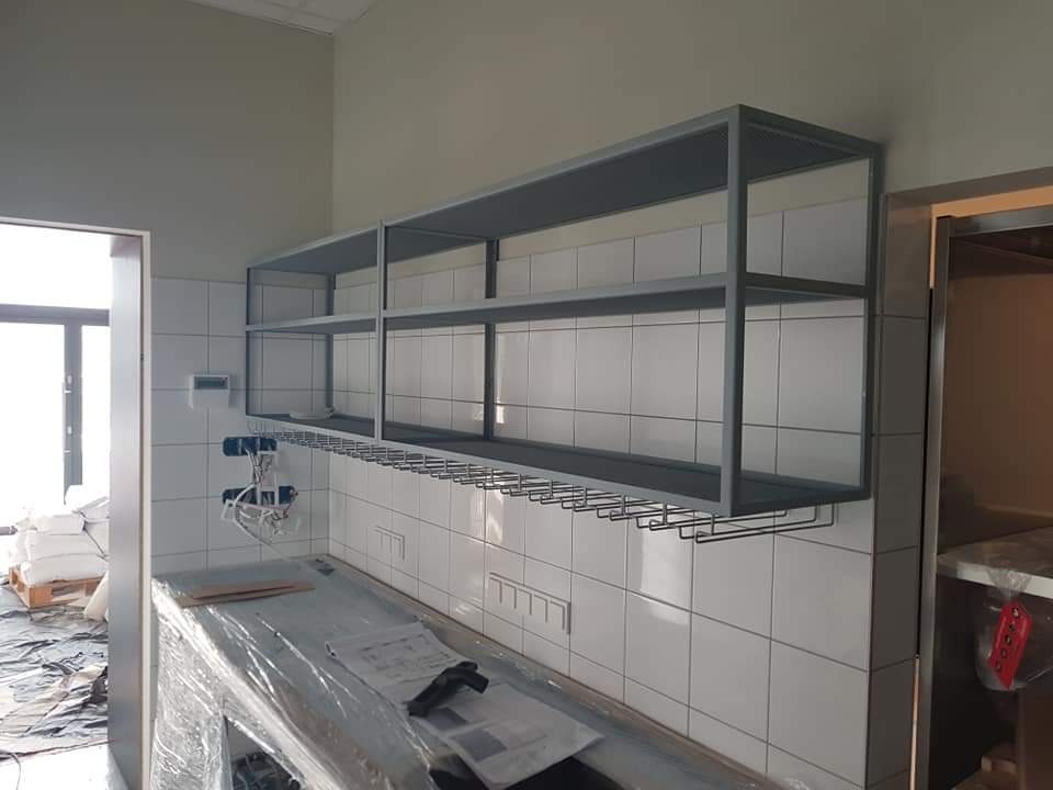 Szare, metalowe półki i uchwyt na kieliszki zamontowane nad blatem wyłożonym białymi płytkami w pomieszczeniu w trakcie wykończenia.