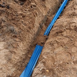 Pro Light Instalacje Elektryczne Łukasz Czajka - Głęboki rów ziemny z ułożoną niebieską osłoną ochronną dla przewodów, otoczony brązową ziemią z kamieniami, widoczny fragment buta.