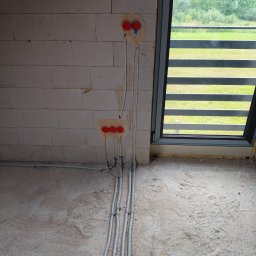 Pro Light Instalacje Elektryczne Łukasz Czajka - Szare peszle z przewodami elektrycznymi ułożone na surowej posadzce i prowadzone do czerwonych puszek w ścianie z jasnych bloczków, obok dużego okna z widokiem na zieleń.