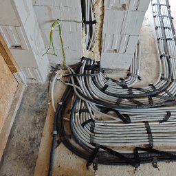 Pro Light Instalacje Elektryczne Łukasz Czajka - Ułożone szare i czarne peszle elektryczne na betonowej posadzce, przygotowane do zalania, z widocznymi przewodami uziemiającymi i pianką izolacyjną w ścianie z białych bloczków.