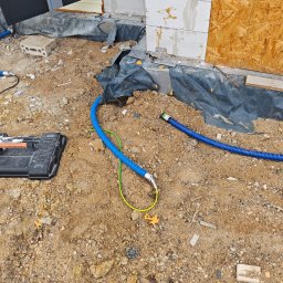 Pro Light Instalacje Elektryczne Łukasz Czajka - Niebieskie peszle z żółto-zielonym przewodem ułożone na piaszczystym gruncie przy fundamencie z białych bloczków, obok czarnej skrzynki narzędziowej.