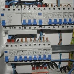 Rozdzielnica elektryczna z zamontowanymi bezpiecznikami Schrack, widoczne kable w peszlach i metalowa szyna montażowa.