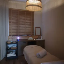 Przytulne wnętrze gabinetu z łóżkiem do zabiegów, rattanową lampą i drewnianymi żaluzjami w oknie.