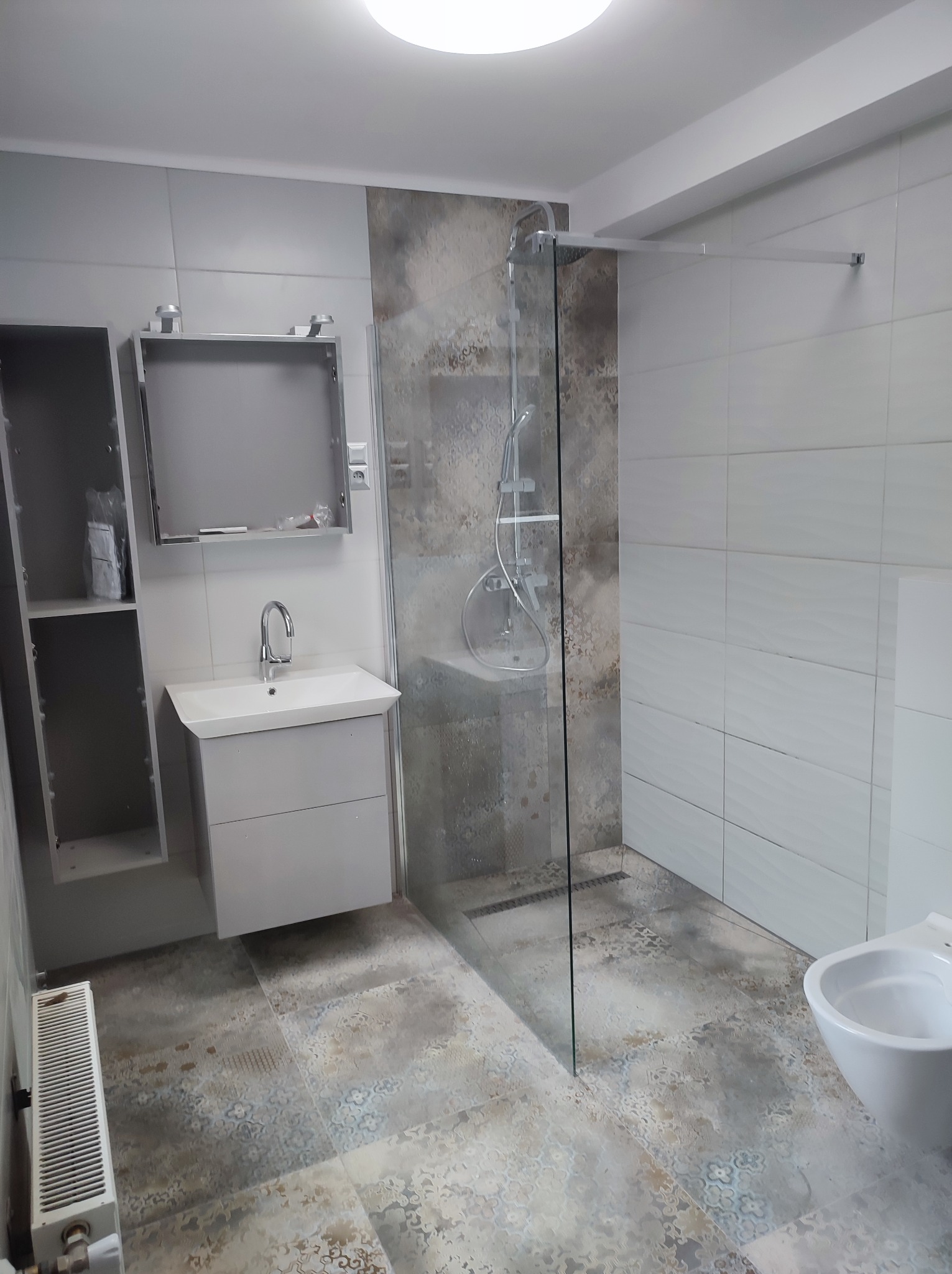 Niedokończona łazienka z kabiną prysznicową typu walk-in, umywalką z szafką, otwartym regałem i grzejnikiem żeberkowym na podłodze w stylu patchwork.