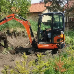 Pomarańczowa mini koparka Kubota KX019-4 w trakcie prac ziemnych w ogrodzie, operator w kabinie, widoczny wykop i pryzma ziemi, w tle dom jednorodzinny i zieleń.