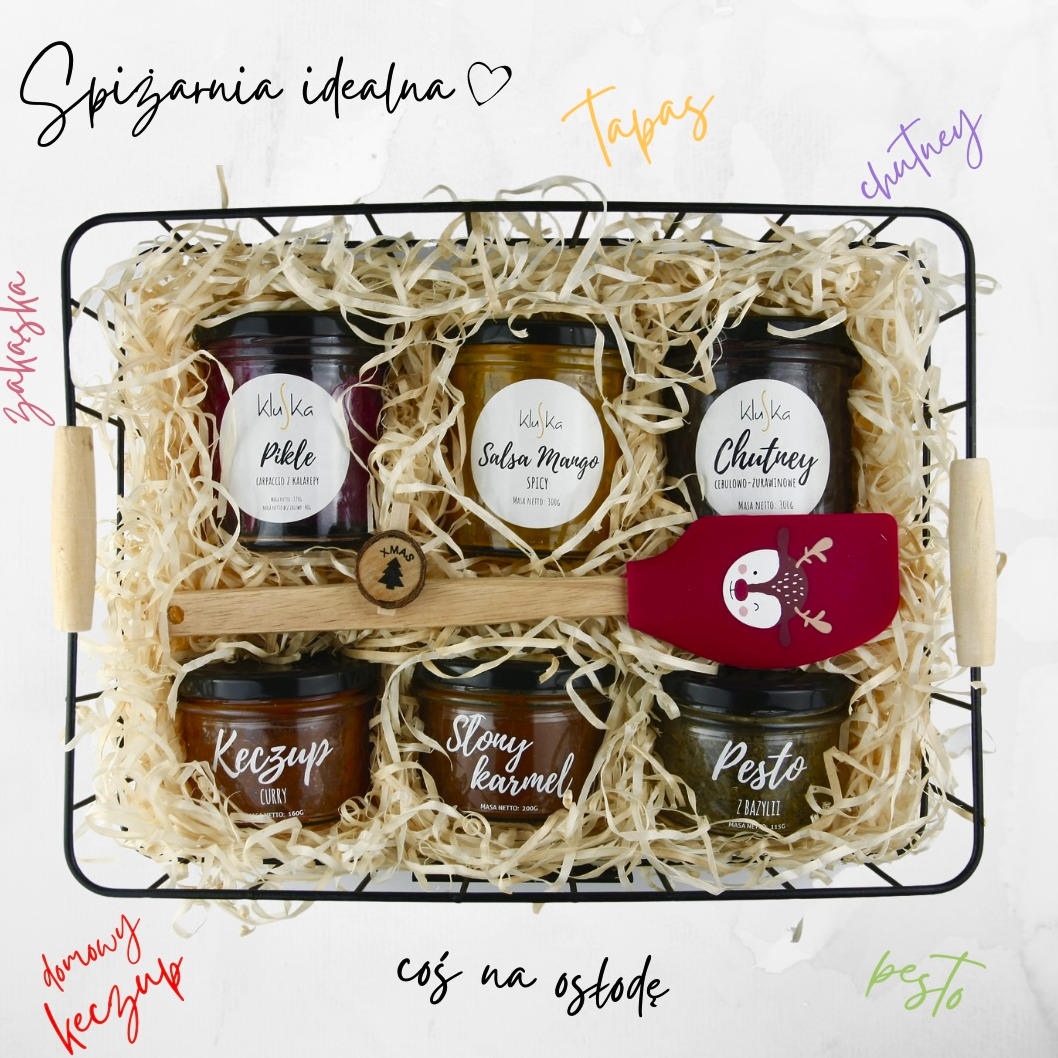 Czarny druciany koszyk wypełniony sześcioma słoikami z przetworami, takimi jak pikle, salsa mango, chutney, keczup curry, słony karmel i pesto, uzupełniony drewnianą szpatułką z dekoracją...