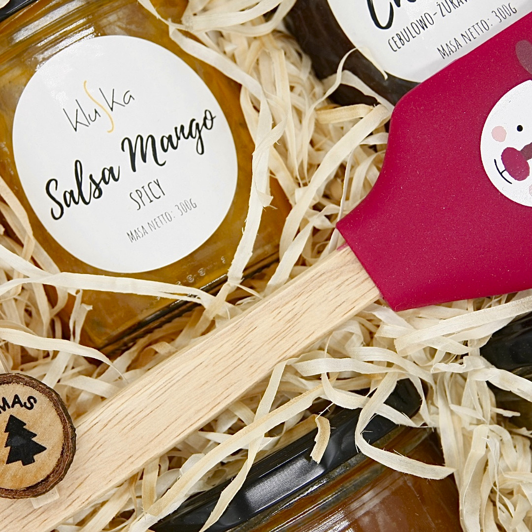 Słoiki z salsą mango i cebulowo-żurawinową w ozdobnym pudełku wypełnionym siankiem, z drewnianą szpatułką z czerwoną silikonową końcówką i małym drewnianym krążkiem z wygrawerowaną choinką.
