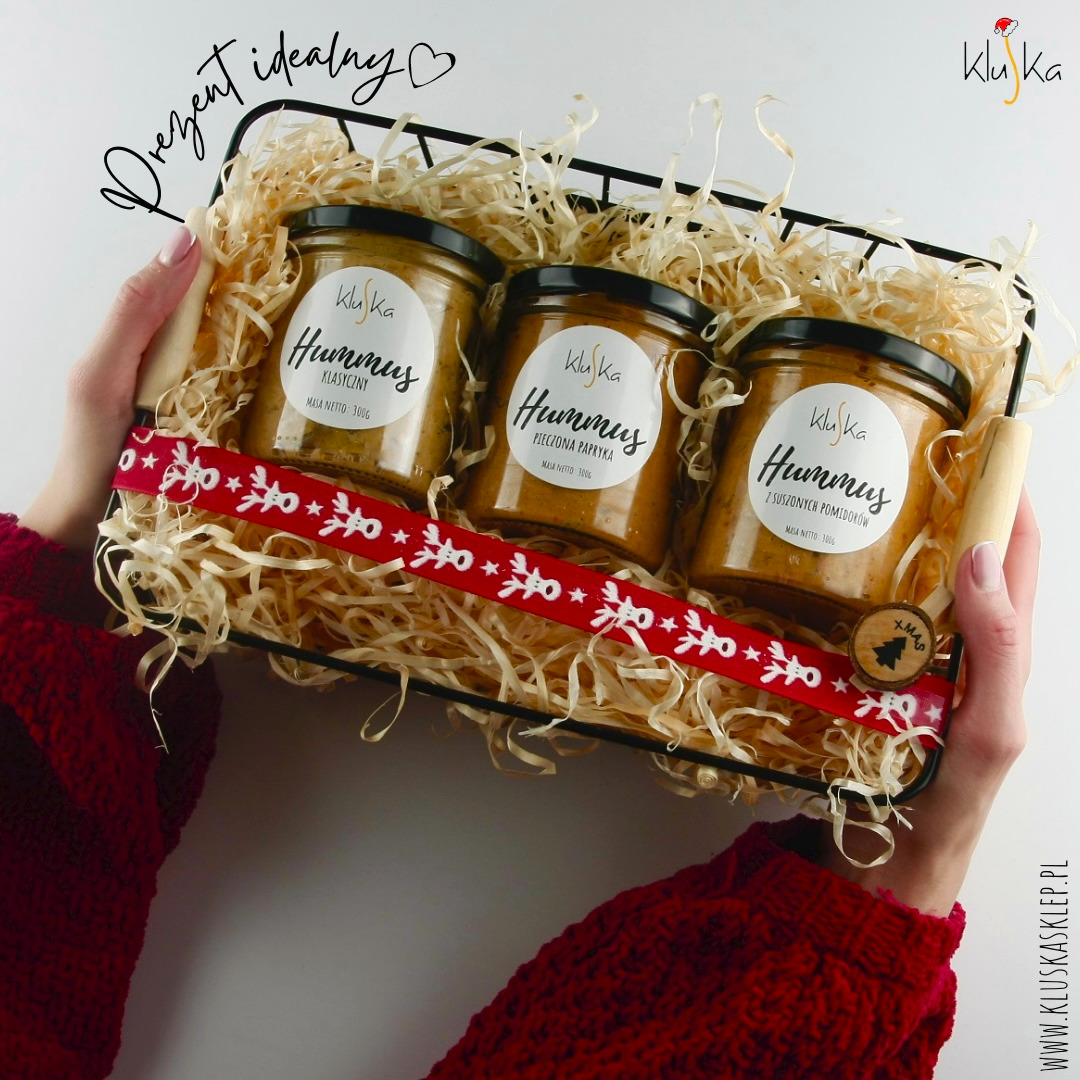 Ręce w czerwonym swetrze trzymają czarny kosz prezentowy wypełniony trzema słoikami hummusu marki Kluska o różnych smakach (klasyczny, pieczona papryka, z suszonych pomidorów) na słomkowym...