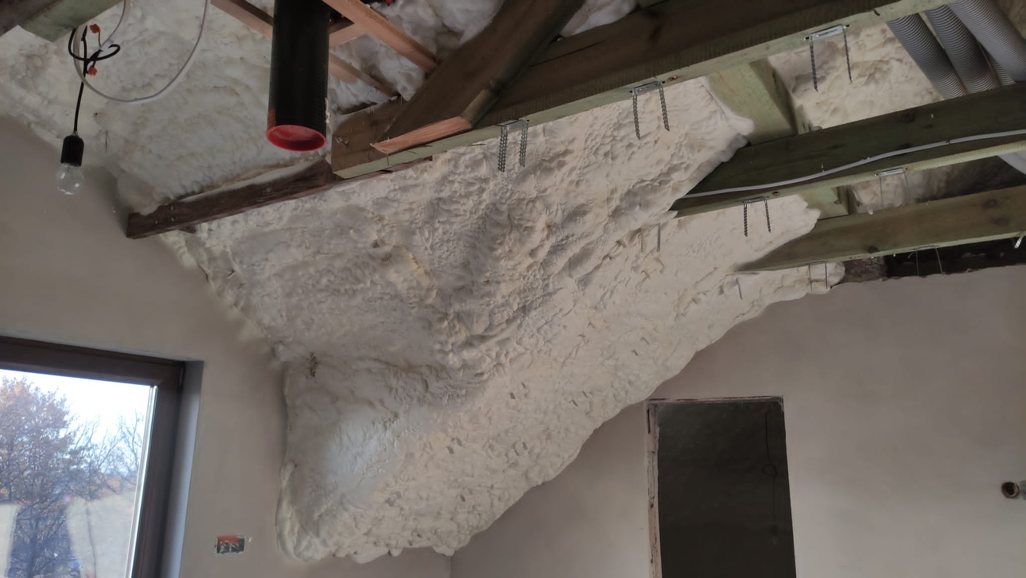 Poddasze z widoczną warstwą izolacyjnej pianki na krokwiach, fragment instalacji wentylacyjnej i widok na okno.