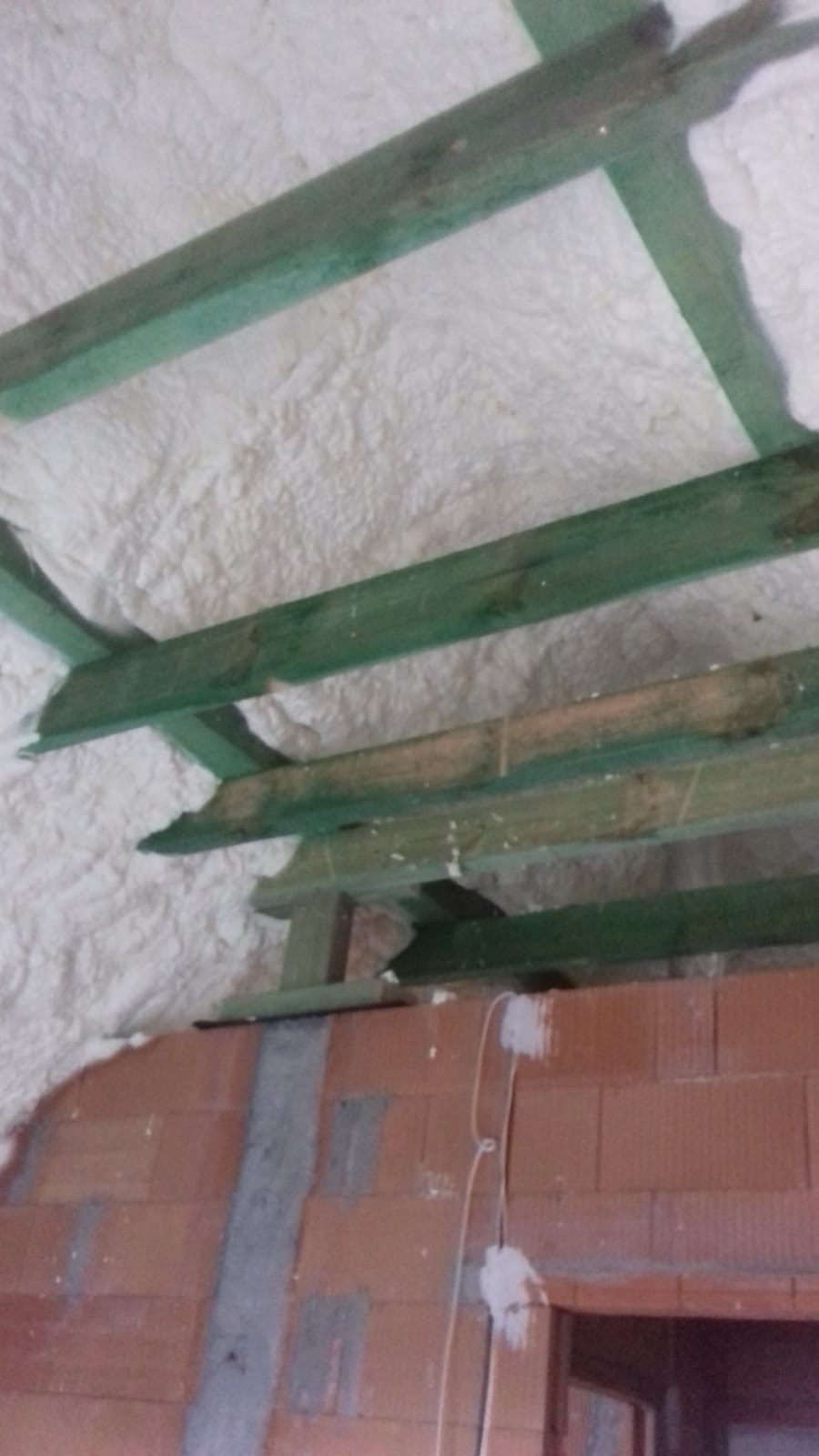 Poddasze z drewnianą konstrukcją więźby dachowej, pokryte warstwą białej pianki izolacyjnej, mur z cegły dziurawki w tle.