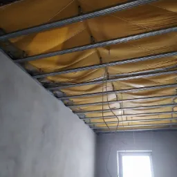 Sufit podwieszany w trakcie montażu: widoczne profile metalowe, żółta folia izolacyjna i wiszące przewody, surowa ściana w tle.