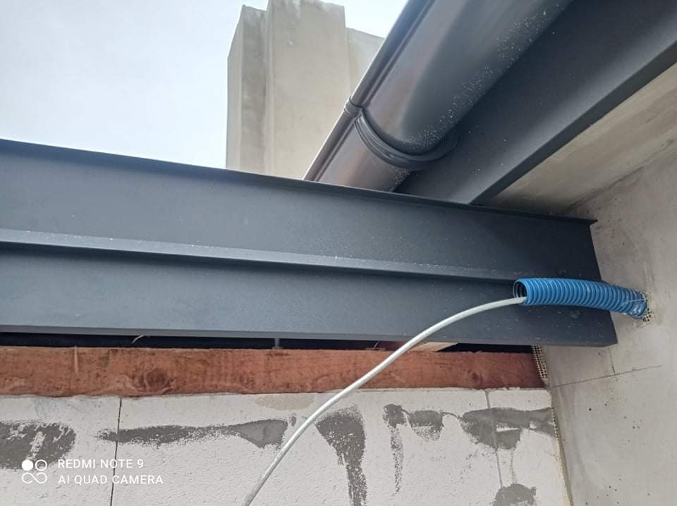 Szara rynna dachowa i stalowa belka wsporcza zamontowane na surowej ścianie z widoczną instalacją elektryczną w niebieskiej rurce karbowanej.