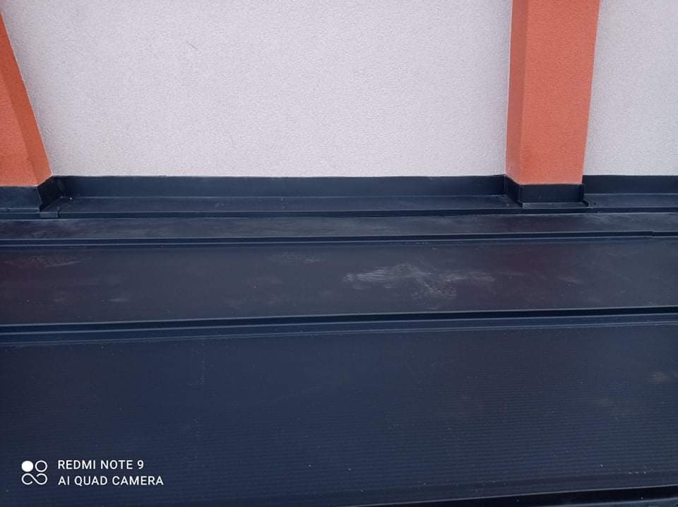 Fragment dachu z blachy trapezowej w kolorze grafitowym, z widocznymi elementami mocującymi do ściany i pomarańczowym słupkiem.