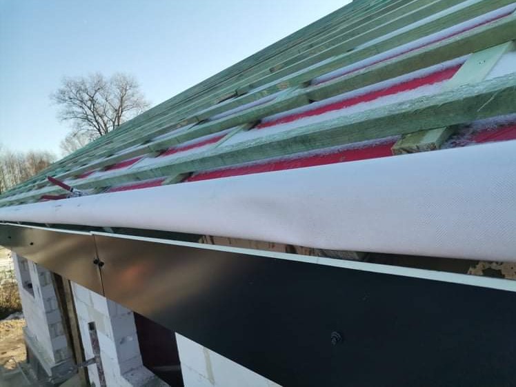 Konstrukcja dachu z drewnianymi belkami, czerwoną membraną i białą folią, widoczne czarne obróbki blacharskie przy krawędzi dachu, budynek w trakcie budowy z białego pustaka.