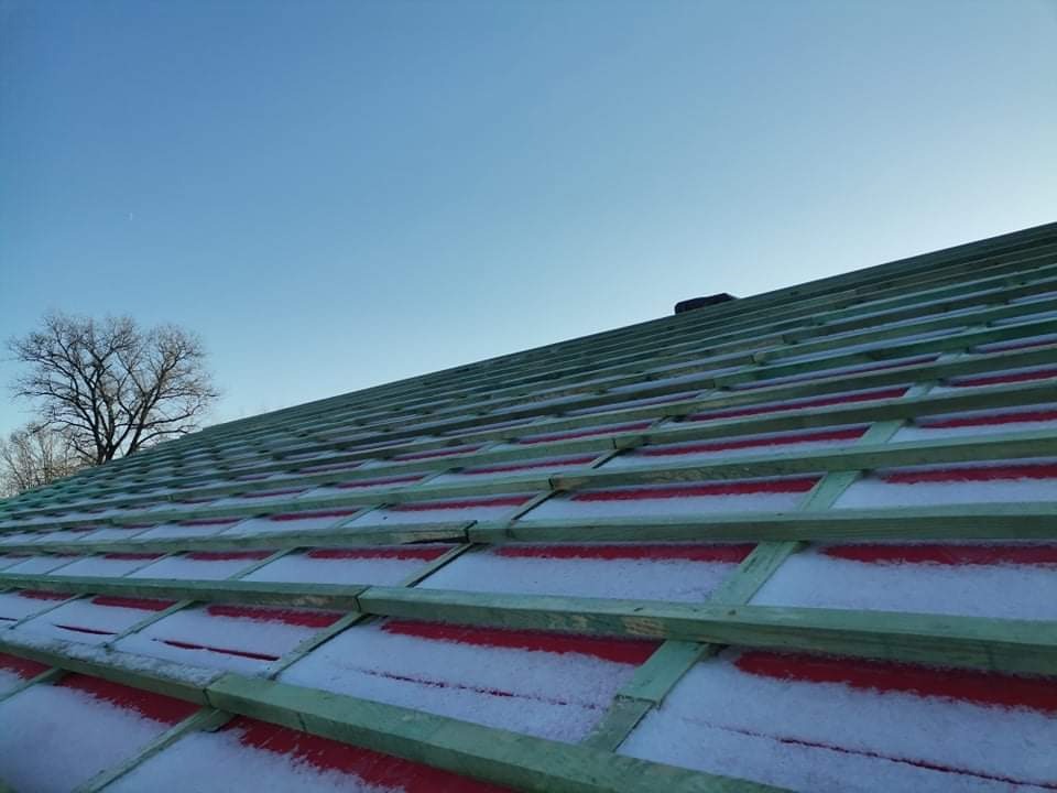 Konstrukcja dachu z drewnianymi łatami i czerwonym podkładem, częściowo pokryta śniegiem, widoczna na tle błękitnego nieba i drzew bez liści.