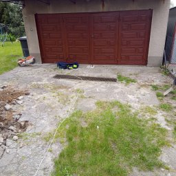 Solis - Roboty Ziemne Instalacje Sanitarne - Prace ziemne przy betonowym podjeździe z kratką ściekową. Widoczny sprzęt budowlany, gruz i ziemia. W tle brama garażowa i ogrodzenie z siatki.