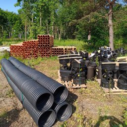 Solis - Roboty Ziemne Instalacje Sanitarne - Różne rodzaje rur kanalizacyjnych (brązowe gładkie, czarne karbowane) i studzienki kanalizacyjne składowane na paletach na terenie budowy, w tle drzewa.