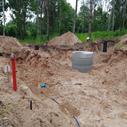 Solis - Roboty Ziemne Instalacje Sanitarne - Wykop z betonowym kręgiem, rurami i czerwoną hydrantową rurą na piaszczystym terenie. W tle drzewa i fragment dachu budynku. Prace ziemne w toku.