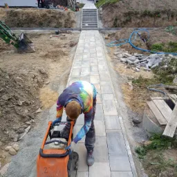 Praca brukarska w Rudzienicach: zagęszczarka w akcji na nowo ułożonej ścieżce z kostki, z mini koparką w tle i schodami prowadzącymi do domu.