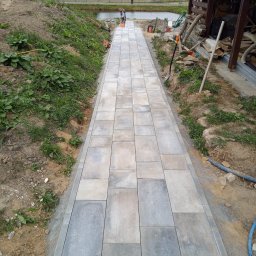 Solis - Roboty Ziemne Instalacje Sanitarne - Ułożona ścieżka z kostki brukowej w ogrodzie, prowadząca w kierunku stawu. Widoczna osoba i narzędzia budowlane, otoczenie naturalne z roślinnością i budynkami.