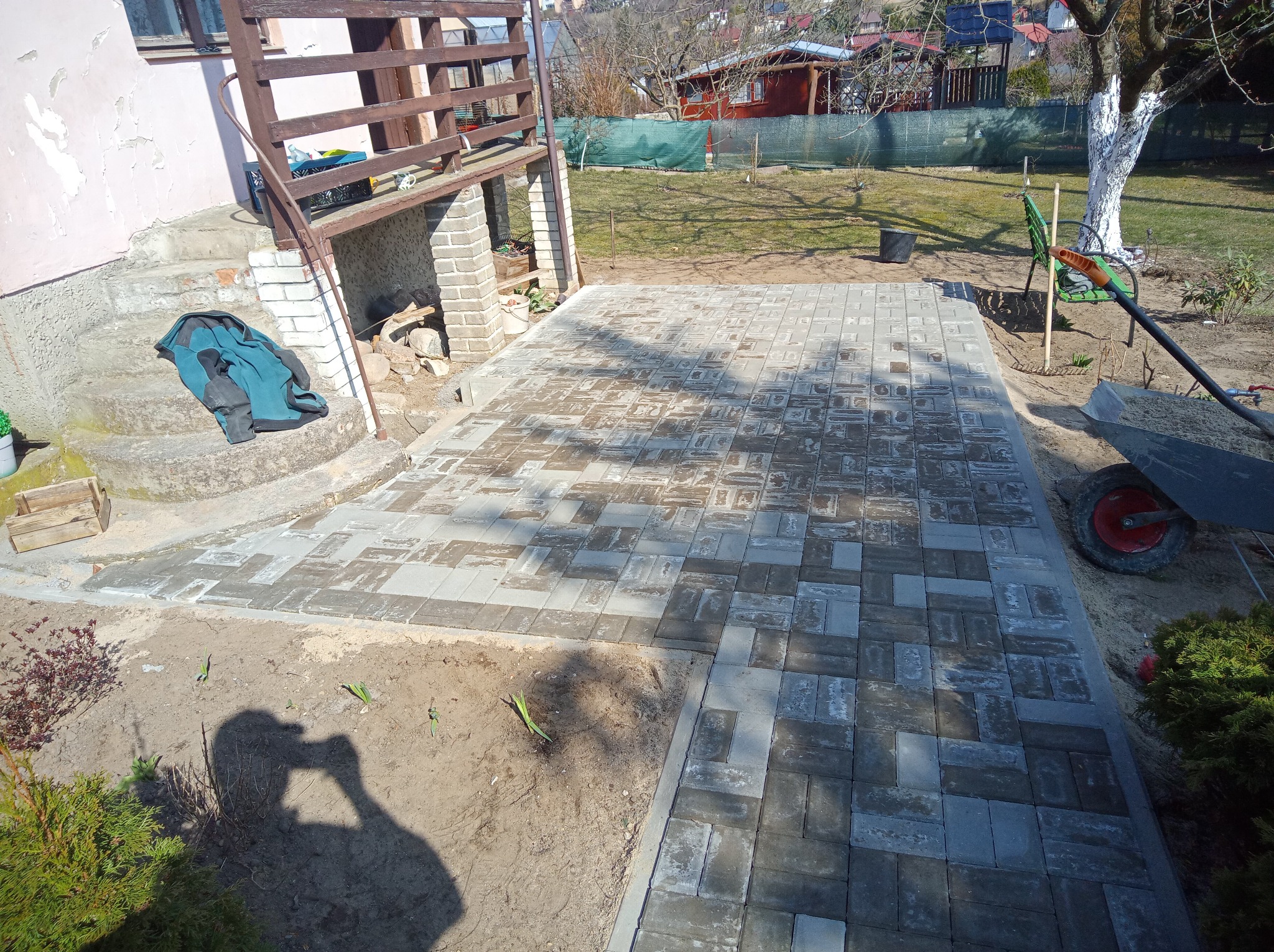 Fragment ułożonej kostki brukowej na posesji, widoczna taczka z piaskiem, cień fotografa. Kostka w odcieniach szarości, ułożona na podwórku obok domu.