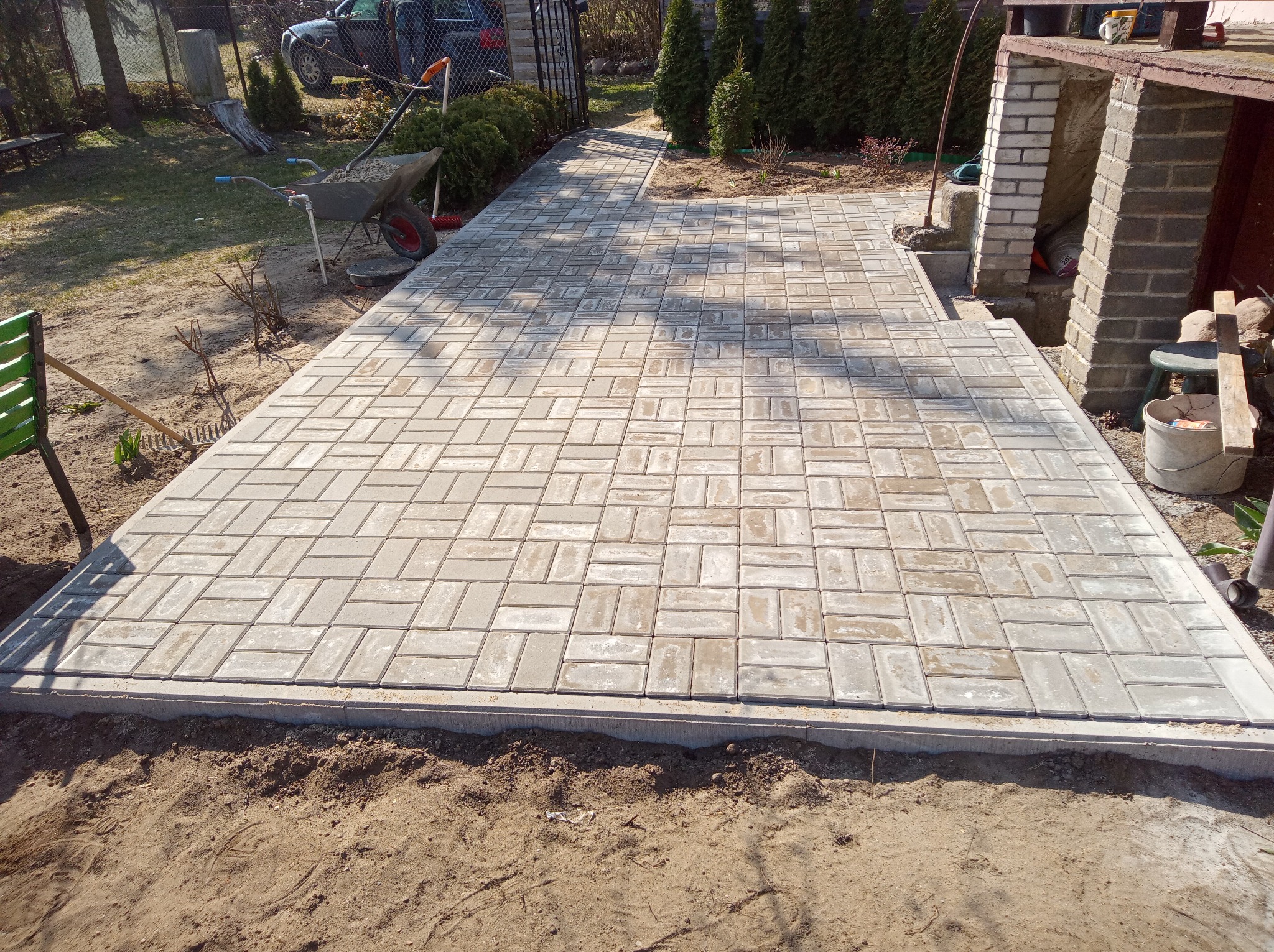 Ułożona kostka brukowa w jodełkę, z widocznym obrzeżem betonowym, na posesji z elementami ogrodu i zabudową. Widok z góry, ciepłe światło słoneczne.