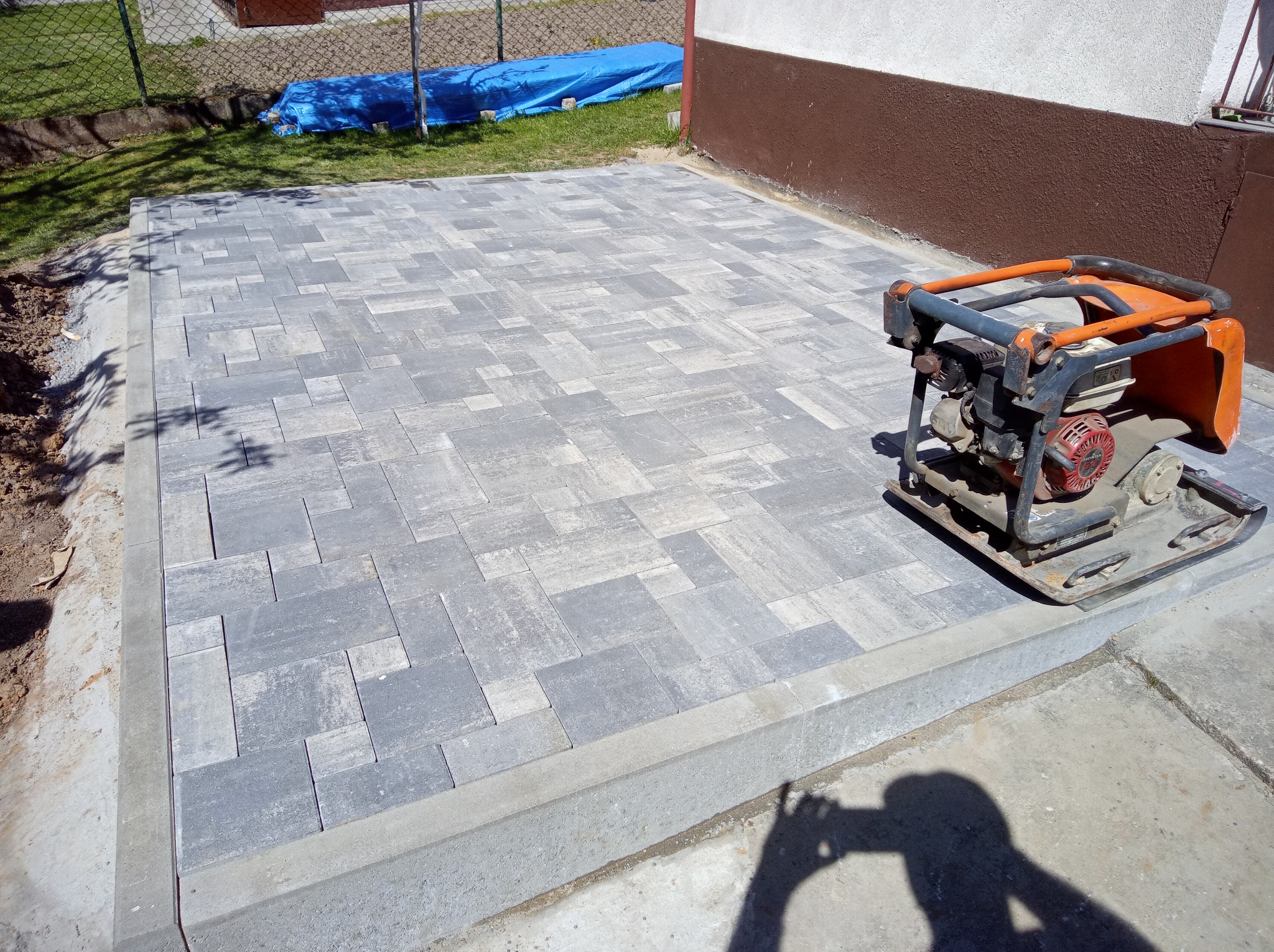 Ułożona kostka brukowa w Rudzienicach, wzór szarych prostokątów, obrzeże betonowe, zagęszczarka obok. Częściowo widoczny trawnik i budynek.