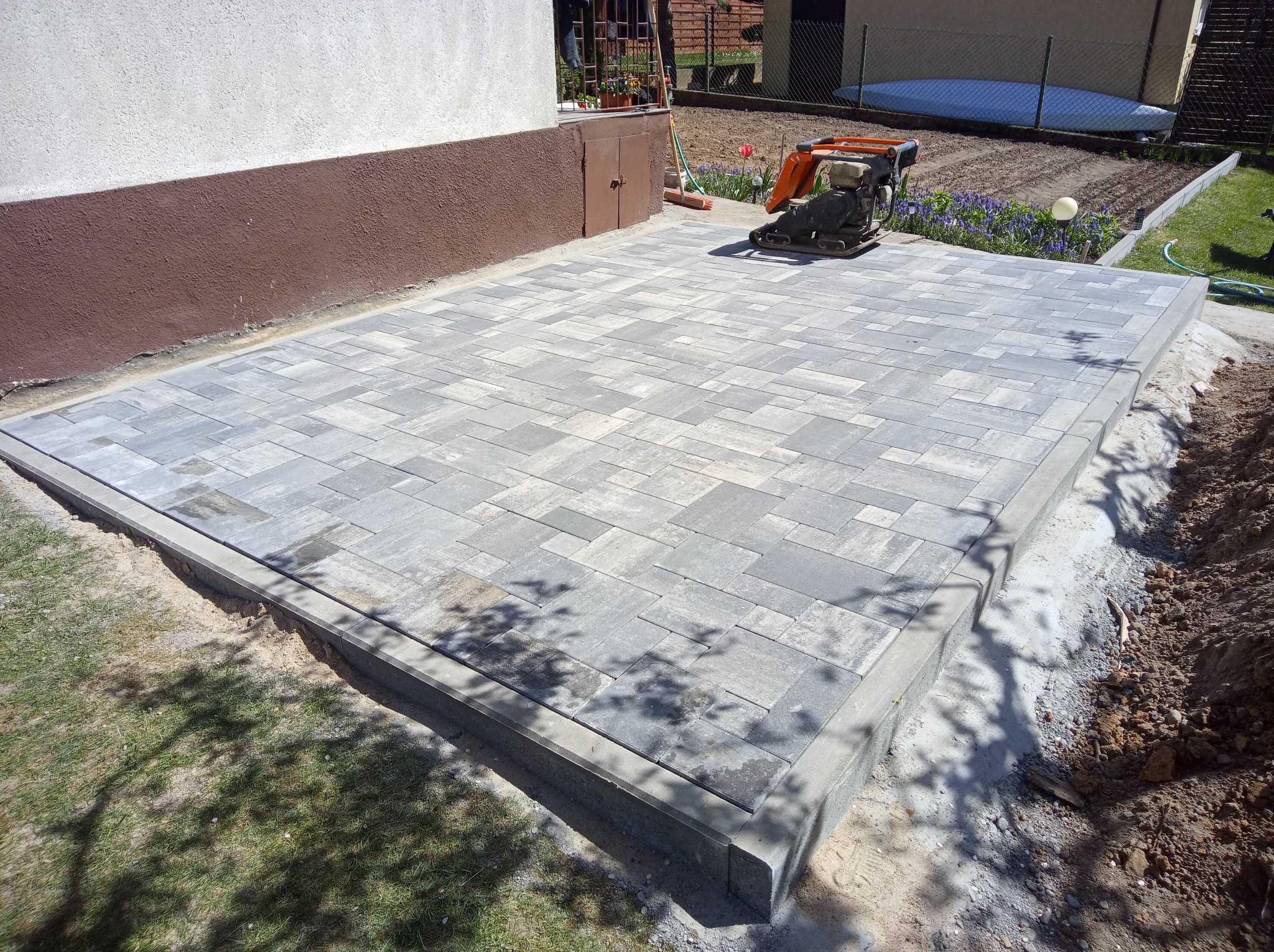 Ułożona, szara kostka brukowa na podwórku, obok budynku. Na kostce stoi zagęszczarka. W tle widać ogródek.