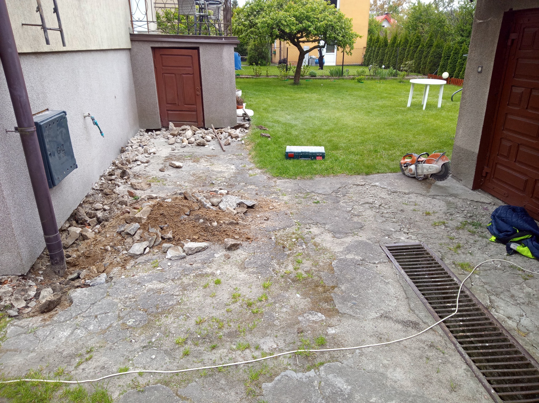 Przygotowanie podłoża: usunięty fragment betonowej nawierzchni, widoczne narzędzia (piła do betonu), skrzynka narzędziowa. Tło: trawnik, budynek.