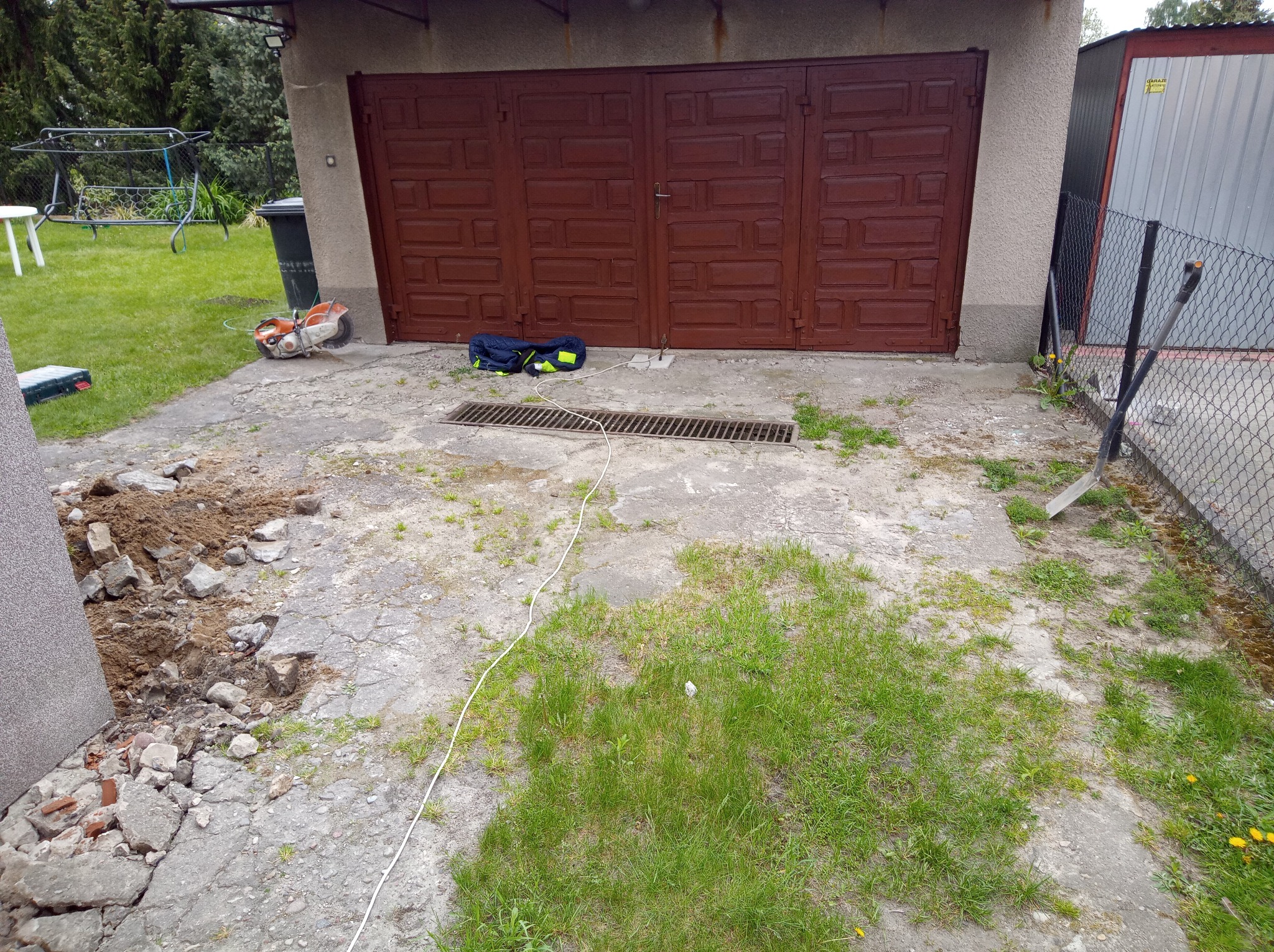 Prace ziemne przy betonowym podjeździe z kratką ściekową. Widoczny sprzęt budowlany, gruz i ziemia. W tle brama garażowa i ogrodzenie z siatki.