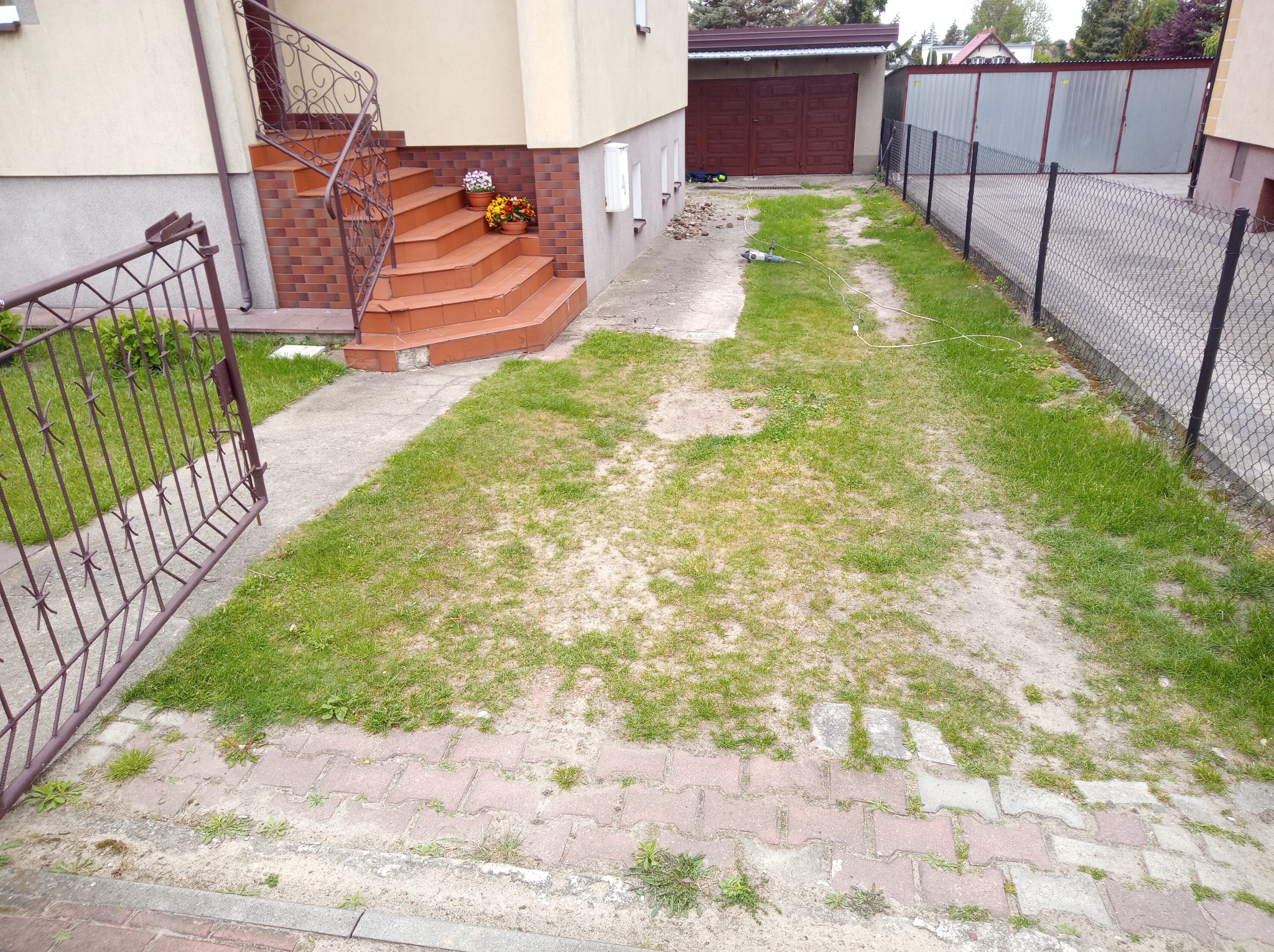Teren przed domem z rozkopaną nawierzchnią, widoczny sprzęt budowlany, sugerujący przygotowanie do prac ziemnych lub układania kostki. Otwarta furtka i schody wejściowe.