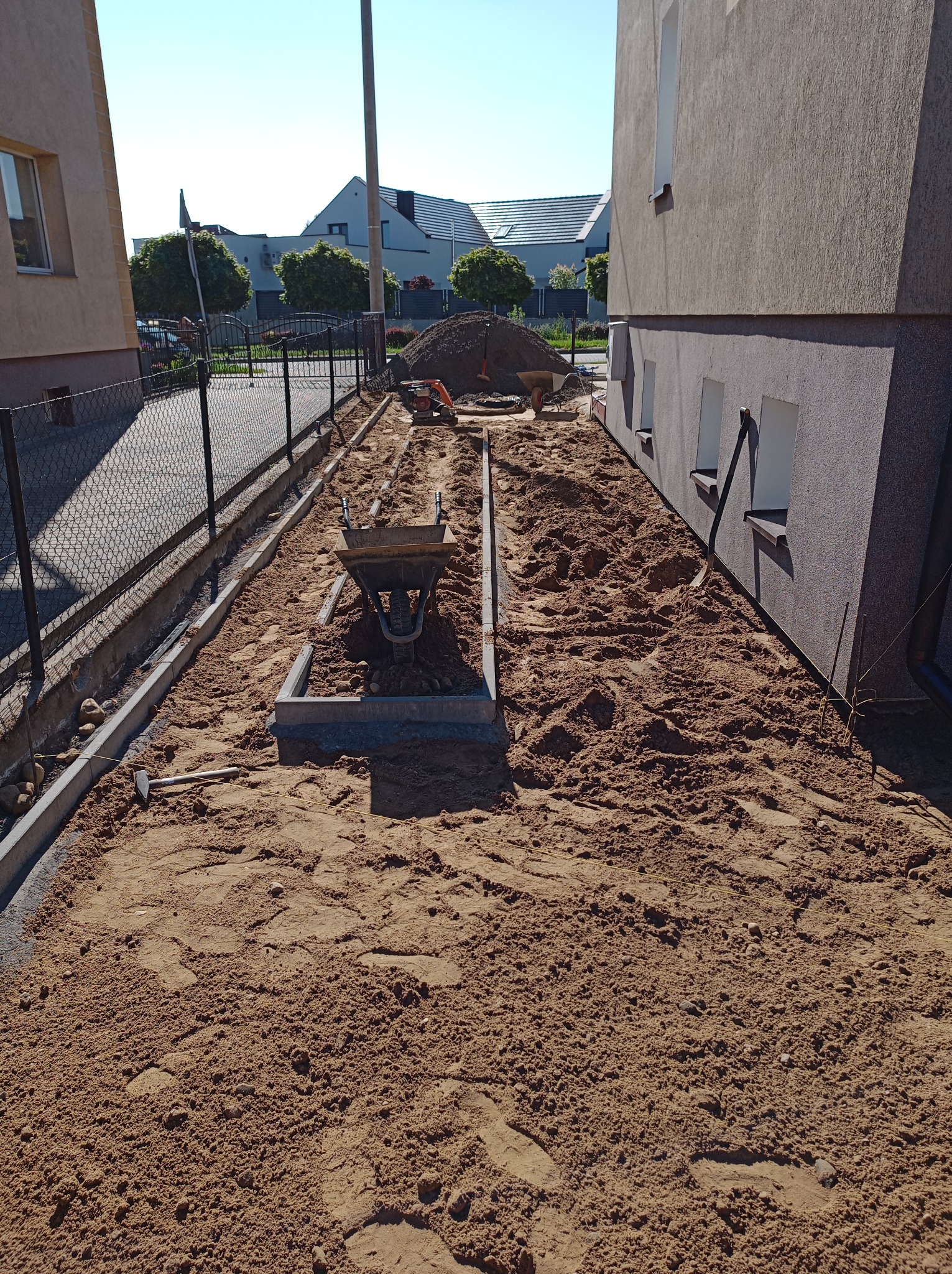 Przygotowanie podłoża pod kostkę brukową: piasek wyrównany, obrzeża ułożone, taczka i ubijarka w tle. Widok z góry na plac budowy między budynkami.