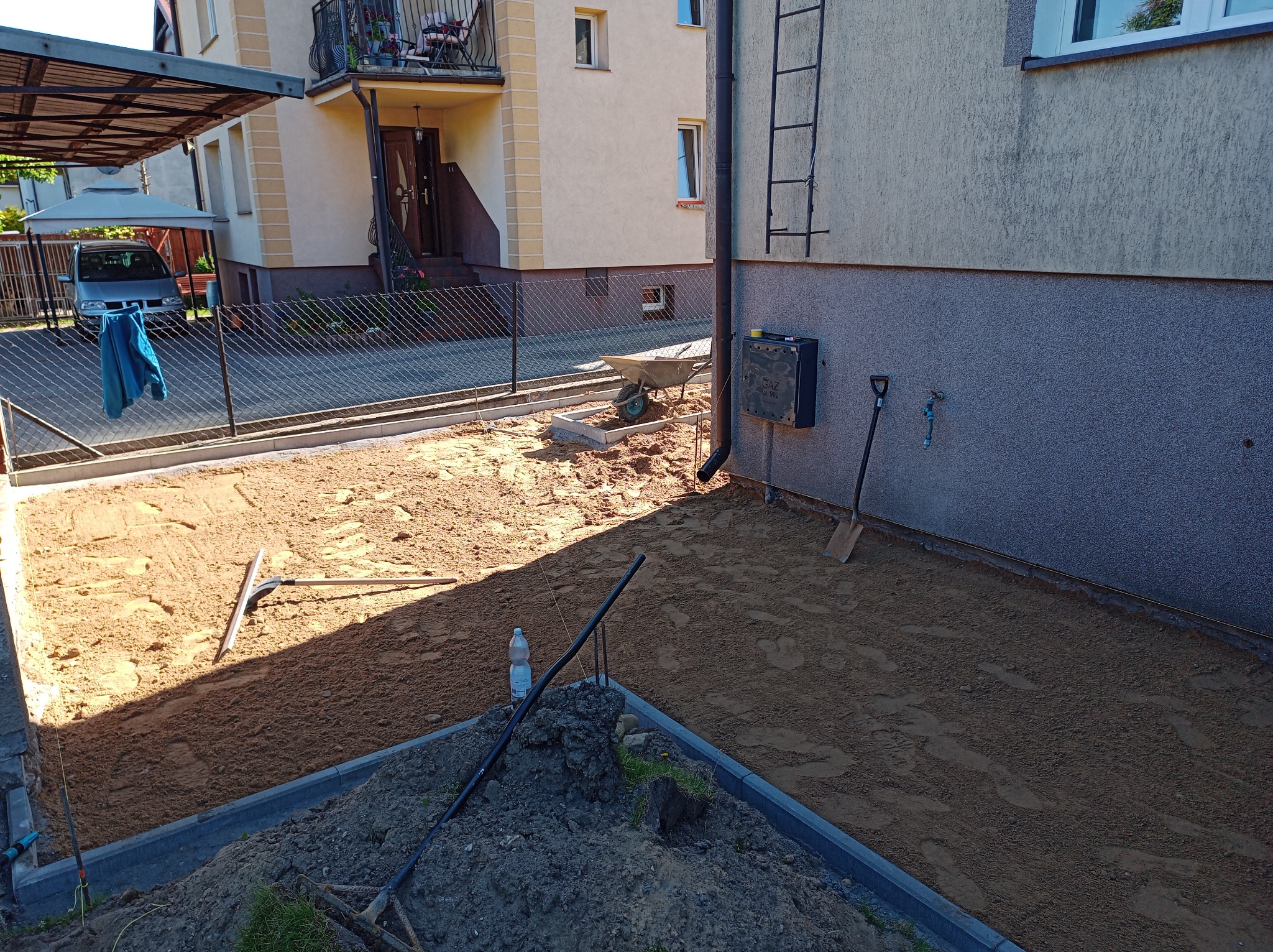 Przygotowanie podłoża pod kostkę brukową: wyrównany piasek, obrzeża ułożone, narzędzia pracy widoczne, dom w tle. Etap prac ziemnych przed ułożeniem kostki.