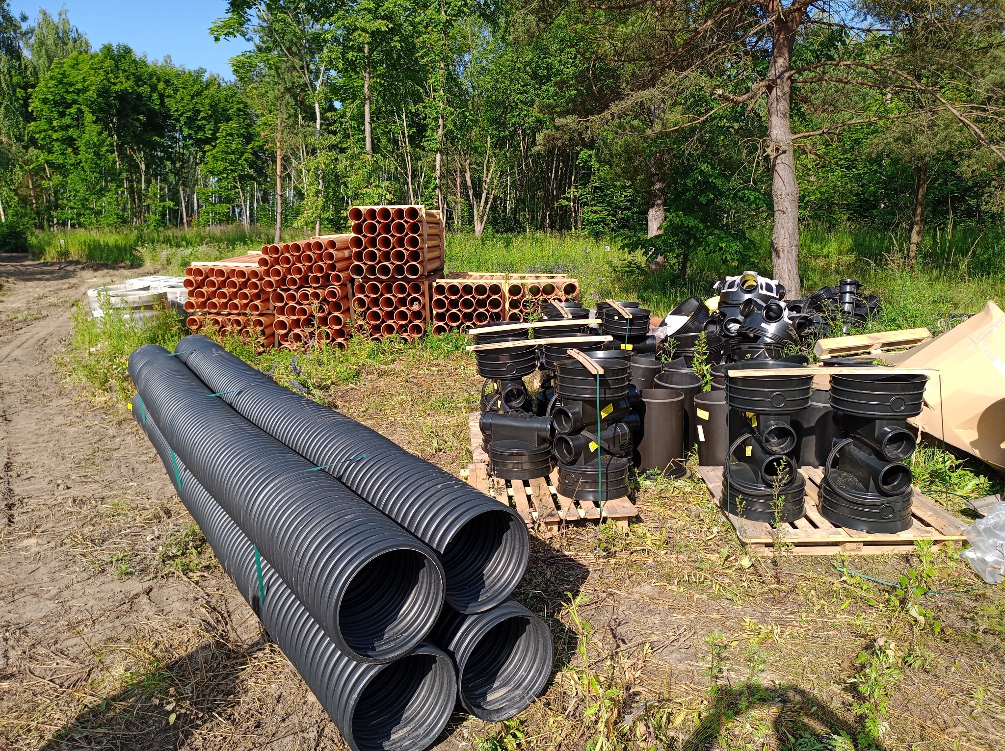 Różne rodzaje rur kanalizacyjnych (brązowe gładkie, czarne karbowane) i studzienki kanalizacyjne składowane na paletach na terenie budowy, w tle drzewa.