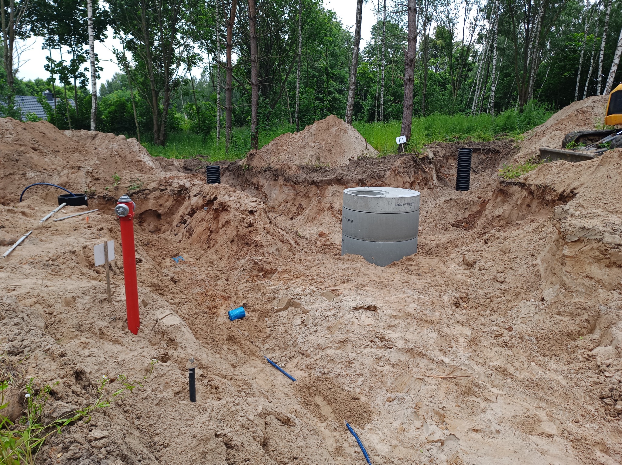 Wykop z betonowym kręgiem, rurami i czerwoną hydrantową rurą na piaszczystym terenie. W tle drzewa i fragment dachu budynku. Prace ziemne w toku.