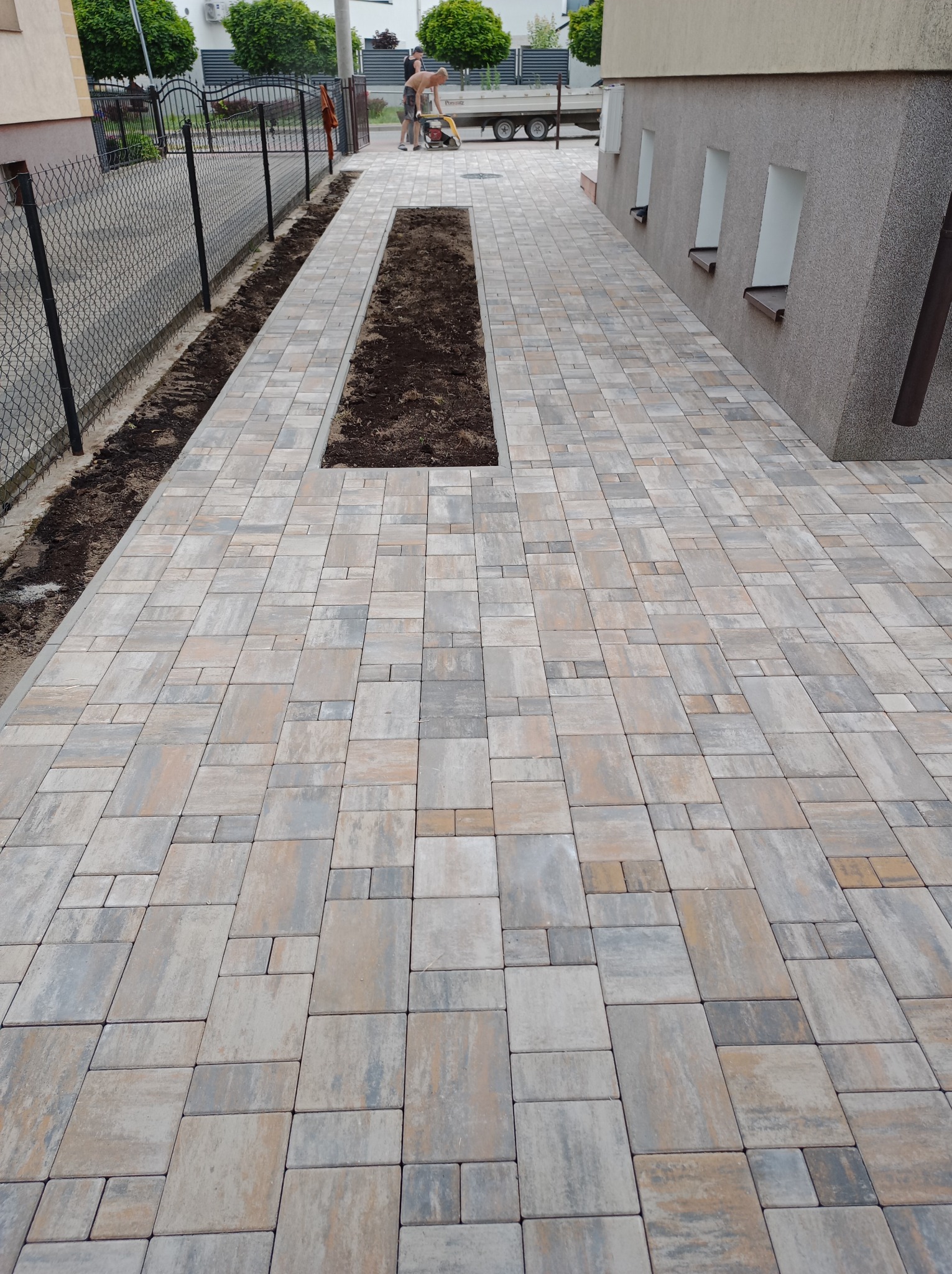 Nowa, wielobarwna kostka brukowa ułożona na podjeździe, z wyznaczonymi miejscami na zieleń i pracownikiem zagęszczającym podłoże w tle.