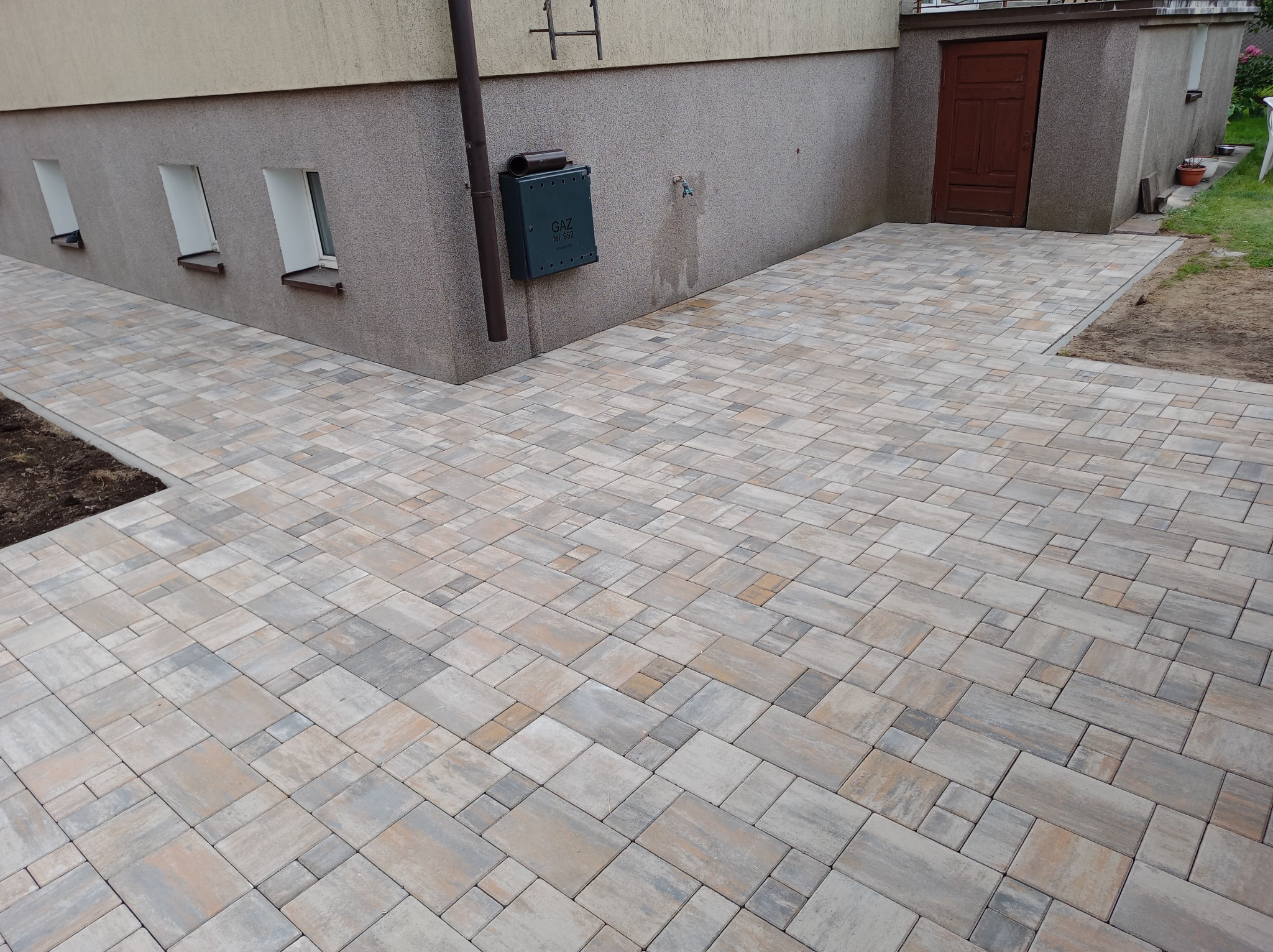 Ułożona kostka brukowa w odcieniach szarości i beżu, tworząca ścieżkę przy budynku z oknami piwnicznymi i zieloną skrzynką gazową. Widoczny fragment trawnika.