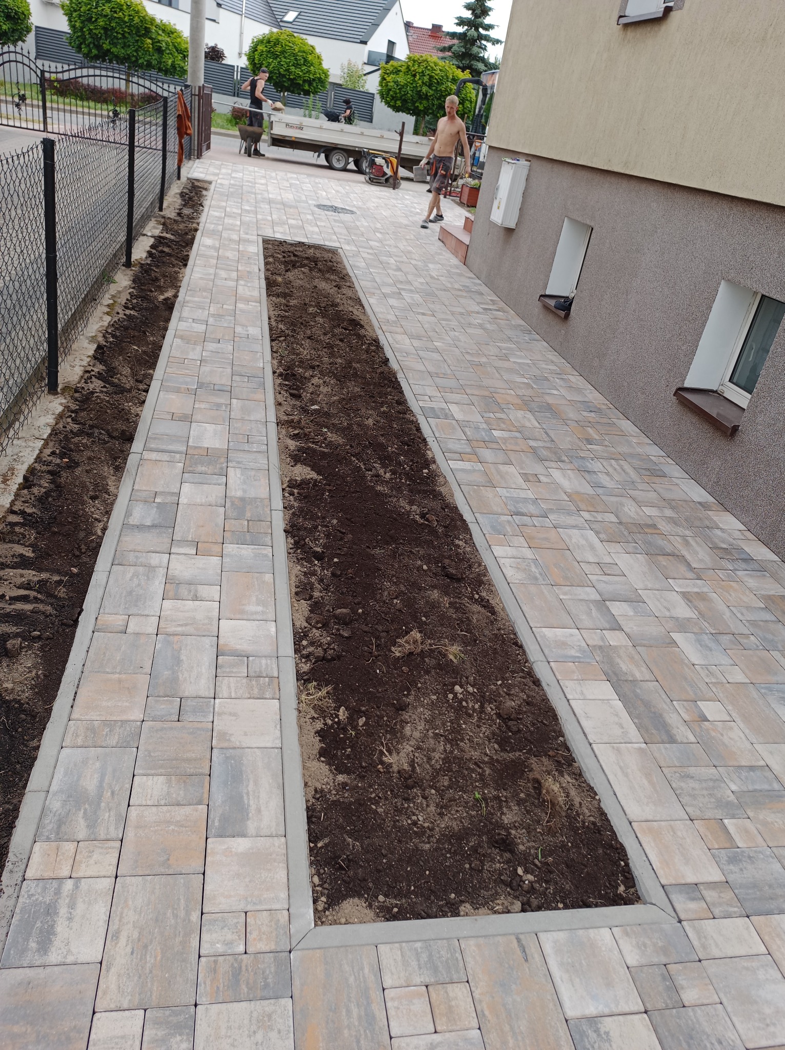 Ścieżka z kostki brukowej w odcieniach szarości i beżu, z wydzielonymi pasami ziemi po bokach i na środku. Prace brukarskie w toku, widoczni pracownicy w tle.