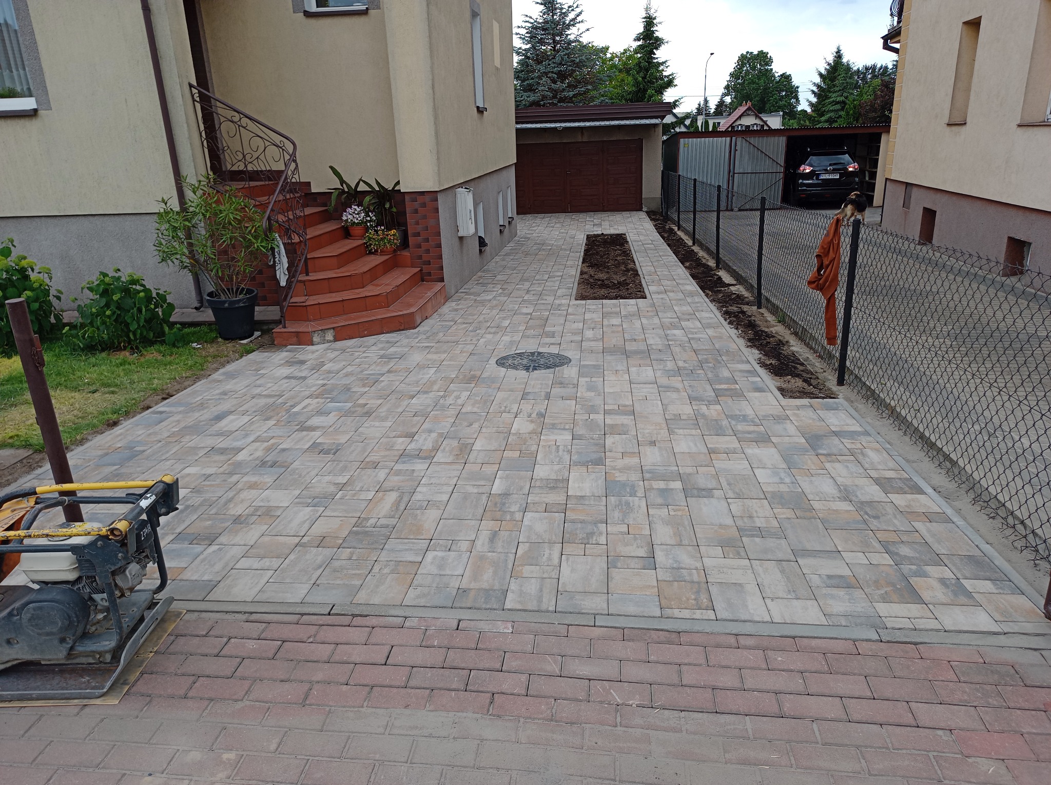 Ułożona kostka brukowa na podjeździe do domu z garażem, widoczna ubijarka do kostki. Część podjazdu z ziemią przygotowana pod nasadzenia, ogrodzenie z siatki.