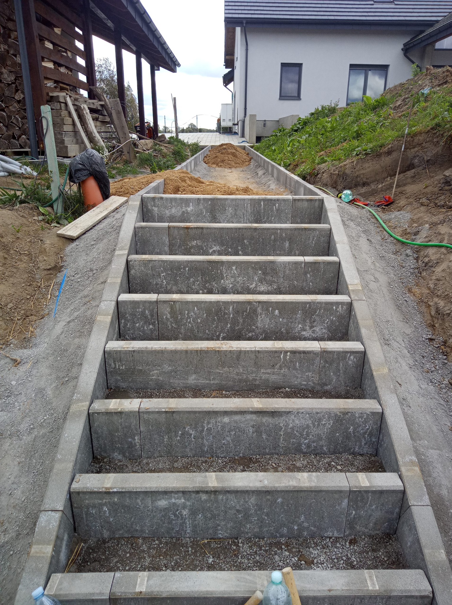 Schody z kostki brukowej w ogrodzie, widok z dołu. Na stopniach rozsypany piasek, w tle dom i sterta drewna. Ujęcie z perspektywy użytkownika schodów.