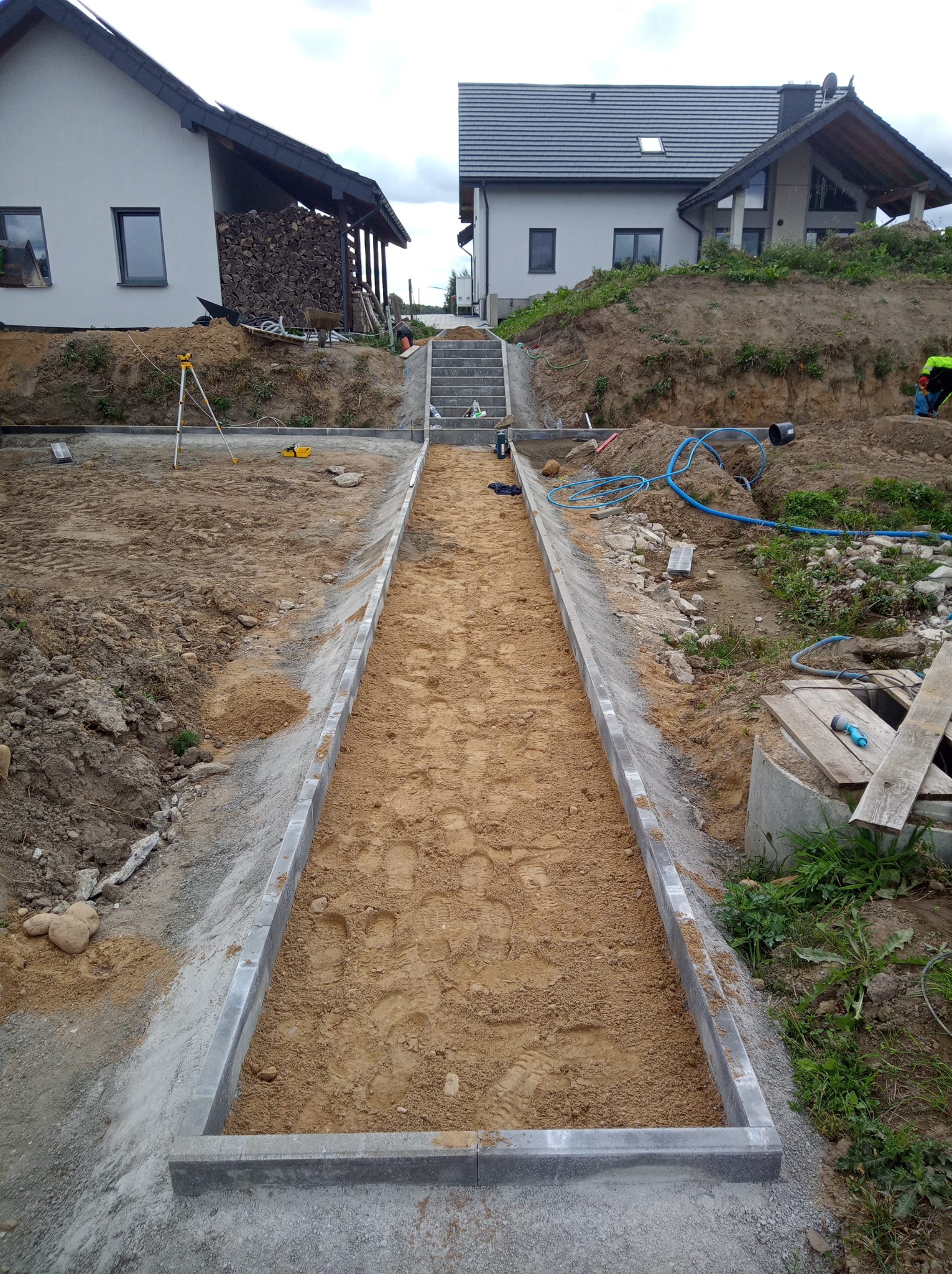 Przygotowanie podłoża pod ścieżkę z kostki brukowej na posesji, widoczne obrzeża i piasek. W tle domy i schody terenowe.