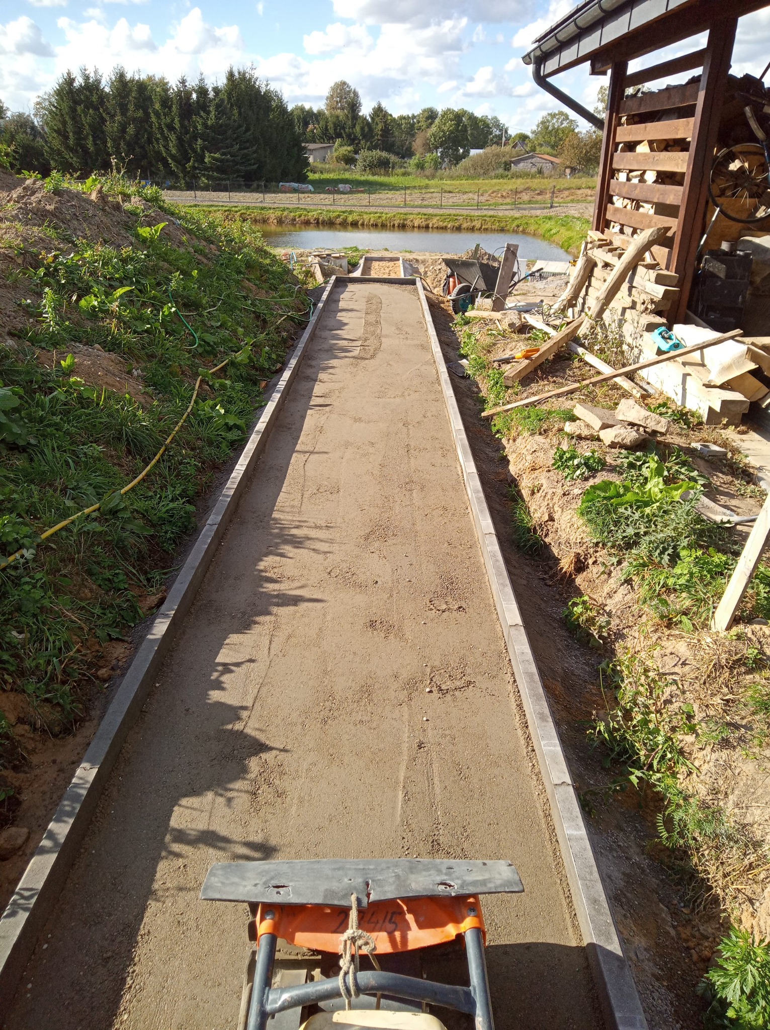 Przygotowanie podłoża pod ścieżkę z kostki brukowej z użyciem zagęszczarki, widok z góry. W tle staw i zabudowania gospodarcze. Ułożone obrzeża.