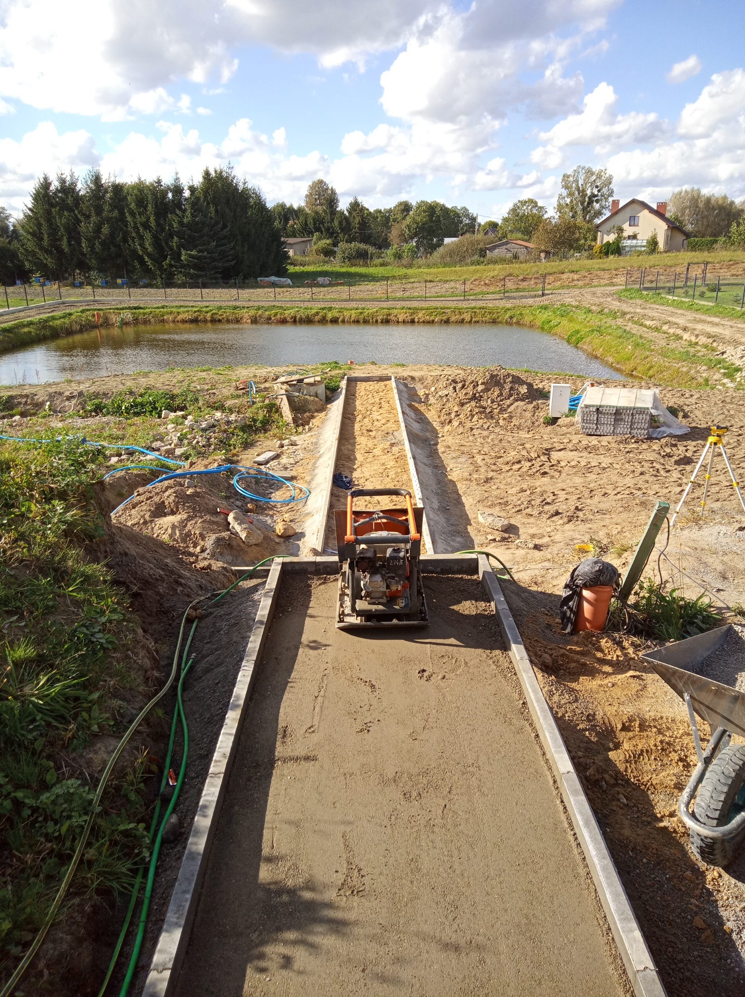 Wyrównywanie podłoża pod kostkę brukową zagęszczarką mechaniczną. Widok na teren budowy z jeziorem w tle, słoneczny dzień.