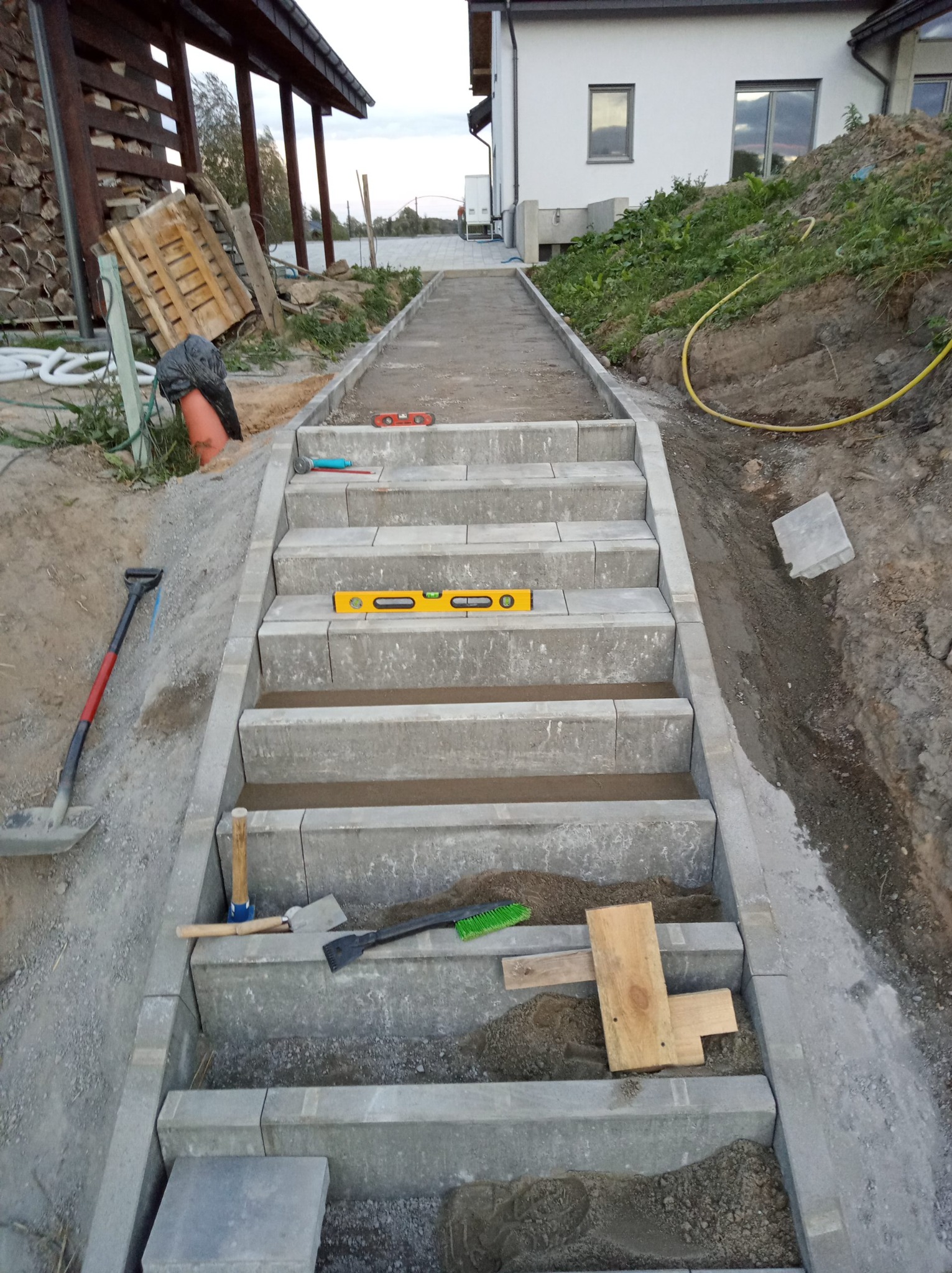 Budowa schodów zewnętrznych z szarej kostki, poziomica na stopniach, łopata i narzędzia obok. Widok z góry na plac budowy z domem w tle.