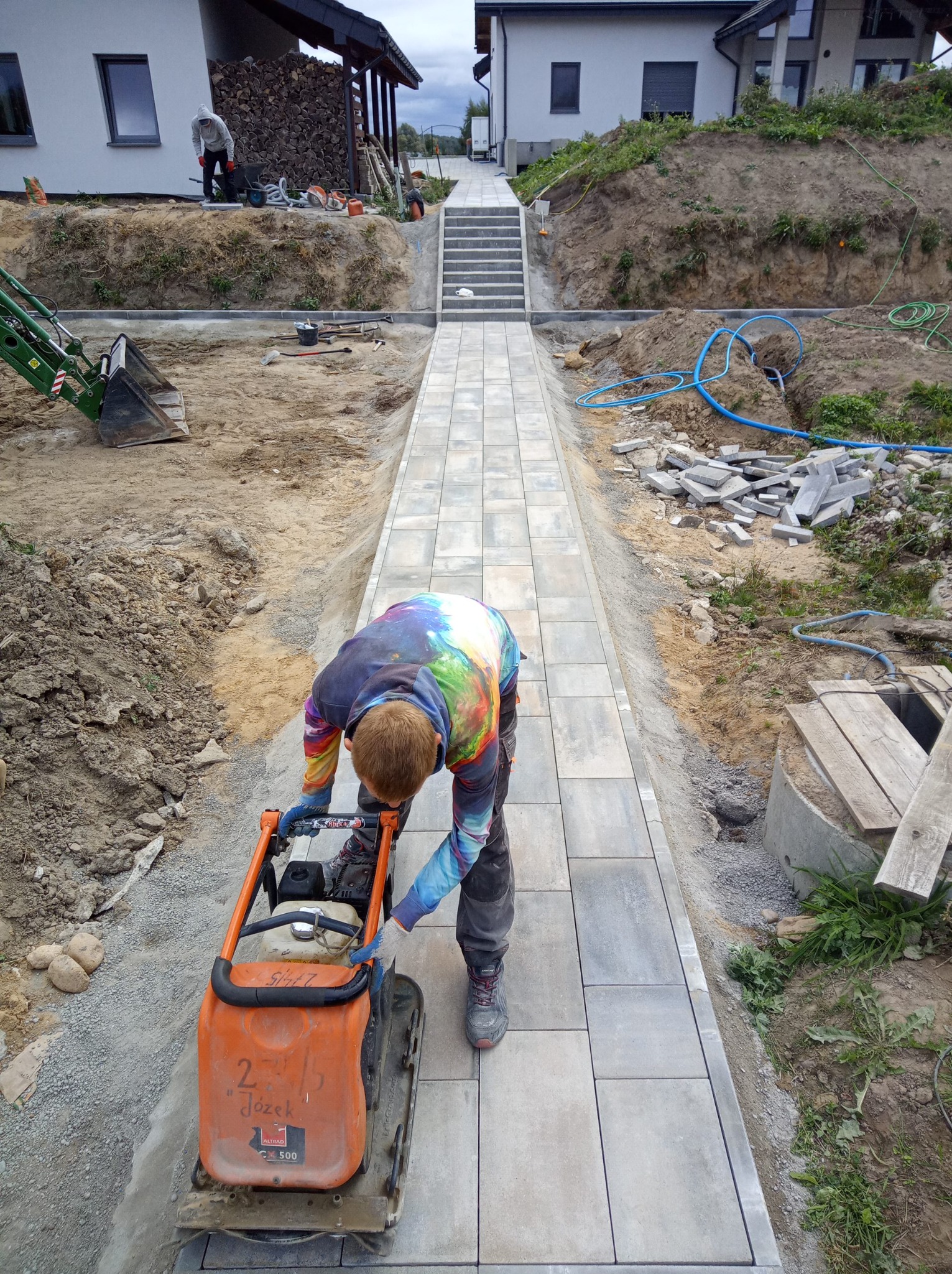Praca brukarska w Rudzienicach: zagęszczarka w akcji na nowo ułożonej ścieżce z kostki, z mini koparką w tle i schodami prowadzącymi do domu.