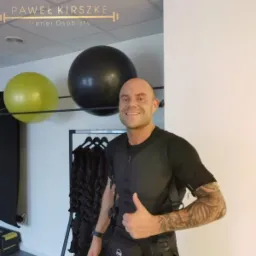 Uśmiechnięty mężczyzna w stroju do elektrostymulacji mięśni, z tatuażem na ramieniu, pokazuje kciuk w górę na tle sali treningowej z piłkami do ćwiczeń i wieszakiem z dodatkowymi kombinezonami.