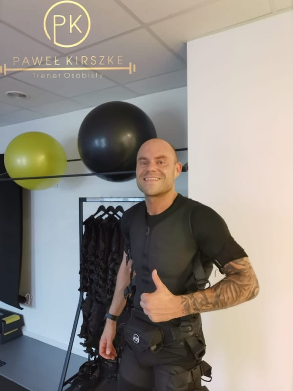 Uśmiechnięty mężczyzna w stroju do elektrostymulacji mięśni, z tatuażem na ramieniu, pokazuje kciuk w górę na tle sali treningowej z piłkami do ćwiczeń i wieszakiem z dodatkowymi kombinezonami.