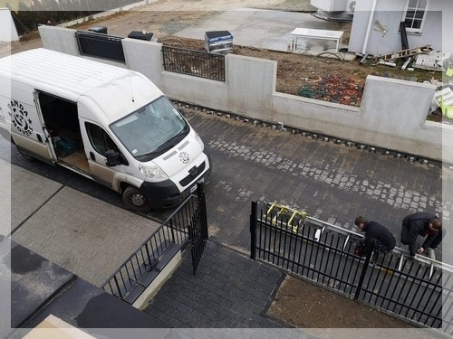 Z góry: biały bus z otwartymi drzwiami zaparkowany na kostce, dwaj mężczyźni przenoszą metalowe przęsło ogrodzeniowe, w tle teren w trakcie prac budowlanych.