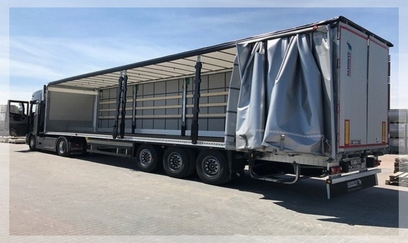 Otwarta naczepa ciężarówki z odsłoniętymi bokami i tylną plandeką, widoczne wewnętrzne zabezpieczenia ładunku, zaparkowana na betonowym placu.