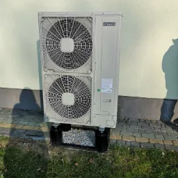 Zewnętrzna jednostka klimatyzacji Hitachi z dwoma wentylatorami, zamontowana na czarnych podstawach wypełnionych białym żwirem, na tle jasnej ściany i trawnika.